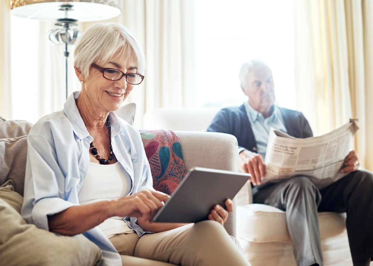 Senior woman on tablet and senior man reading newspaper in living room